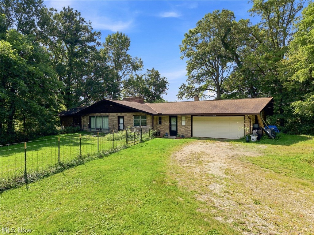 Single-story brick house with an attached garage, gravel driveway, and fenced lawn beneath mature trees.