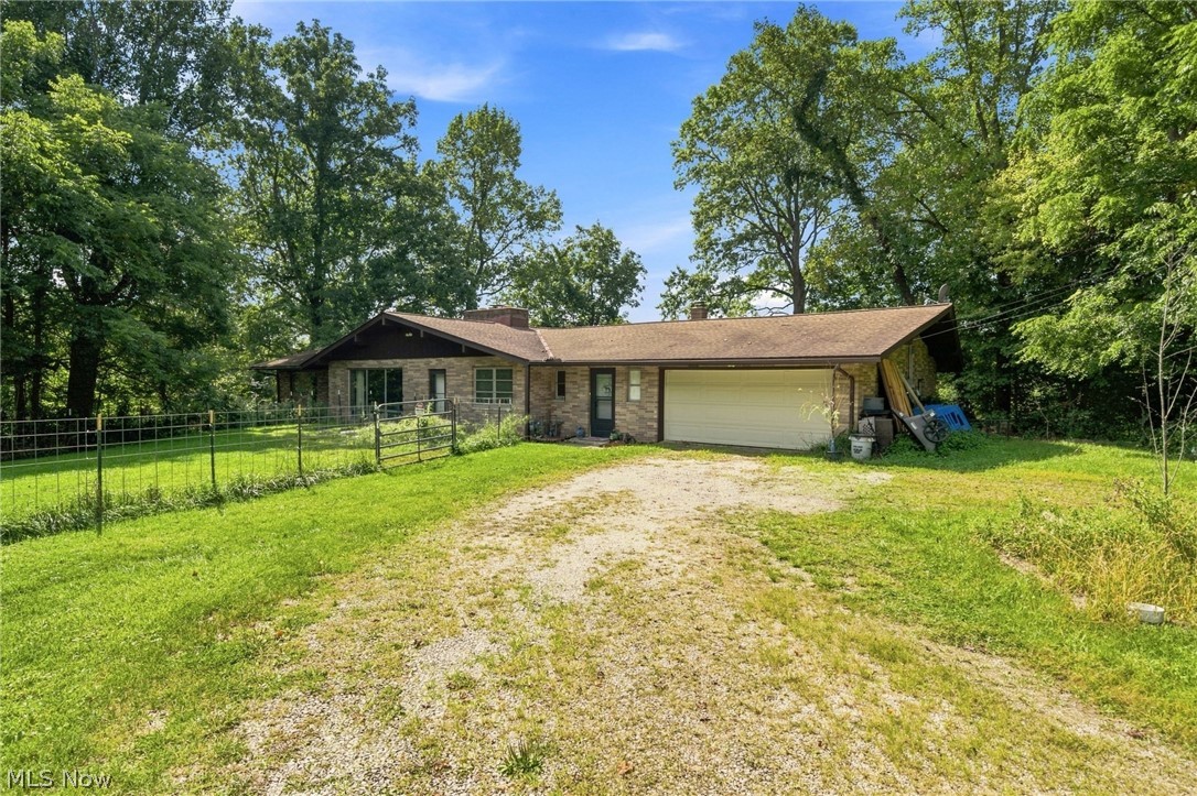 Brick ranch home with an attached garage, gravel driveway, fenced lawn, and mature trees.