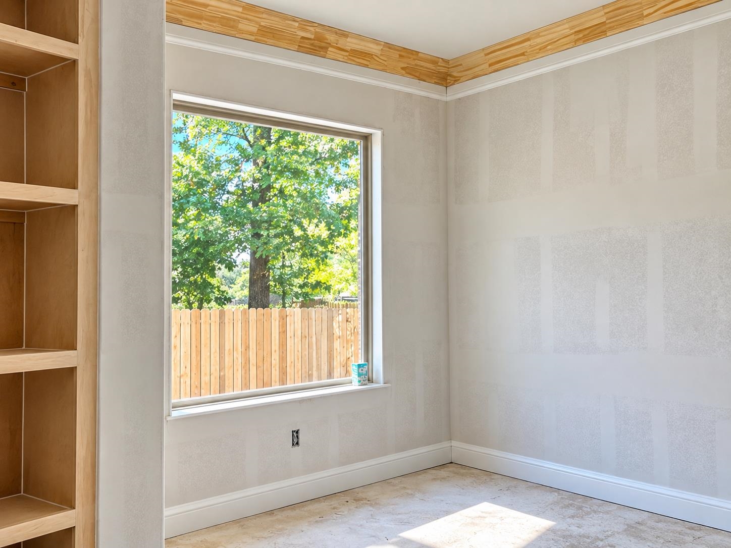 Empty room with built-in wooden shelves, decorative wood crown molding, and a large window overlooking a fenced yard.