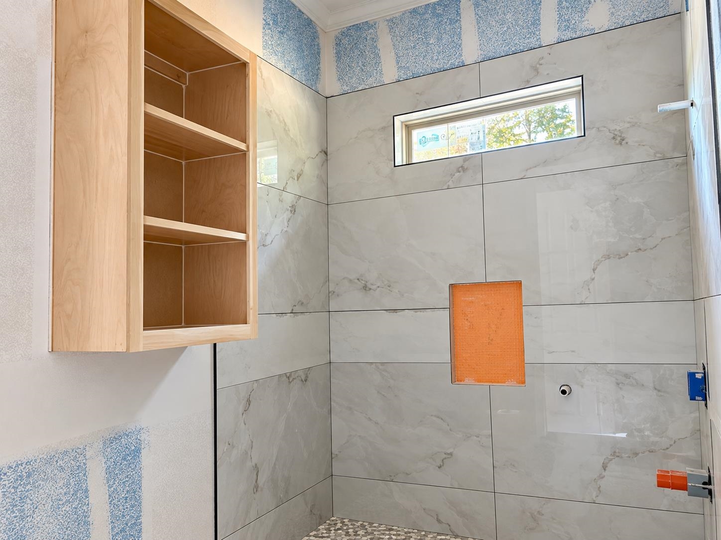 Shower with large marble-look tiles, built-in wood shelves, and a narrow high window.