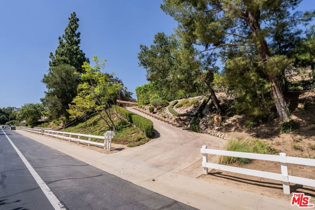 Gated-property driveway entrance with white rail fences, landscaped slopes, and mature trees.