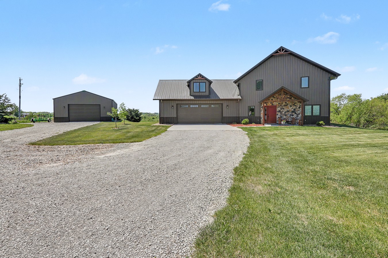 Gray two-story home with a stone entry, red door, gravel driveway, and detached garage.