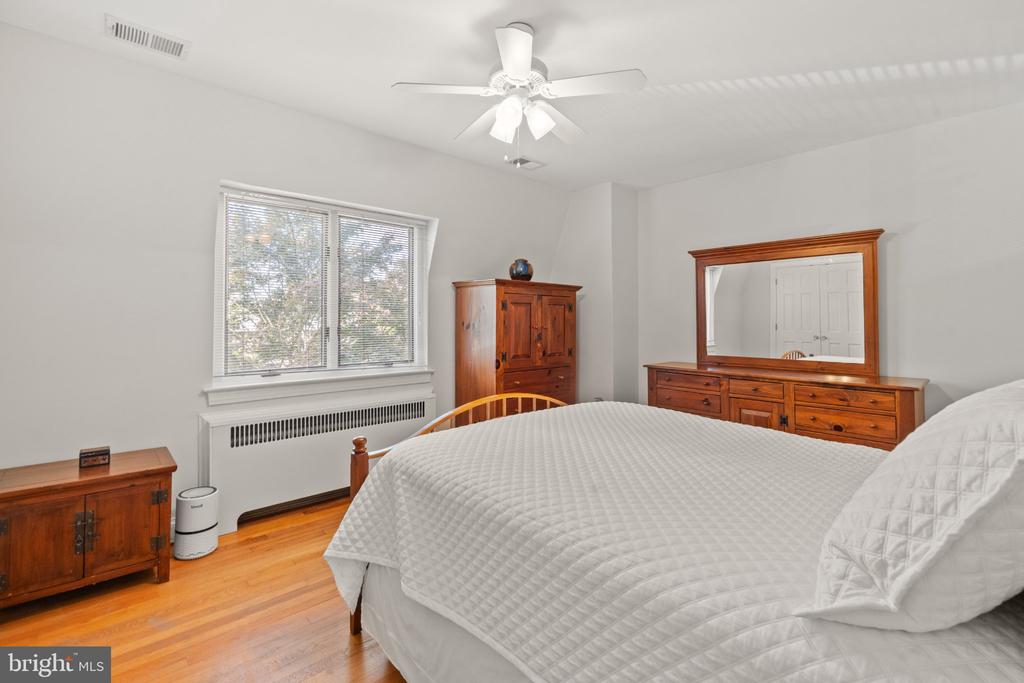 Furnished bedroom with a large white-quilted bed, hardwood floors, and wooden dresser with mirror.