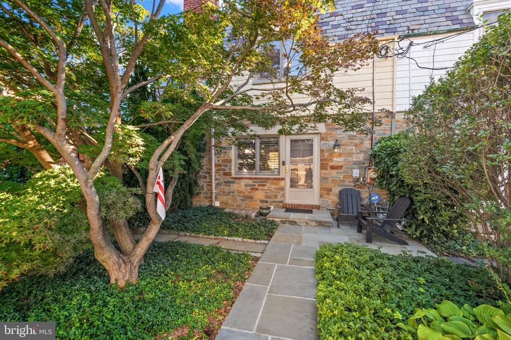 Stone-sided home entrance with a flagstone walkway, leafy garden, and two Adirondack chairs.