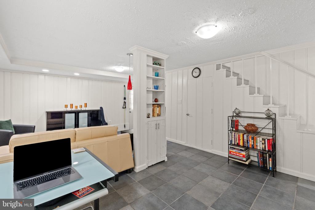 Finished basement living area with a tan sectional, gray tile floor, built-in shelves, and staircase.
