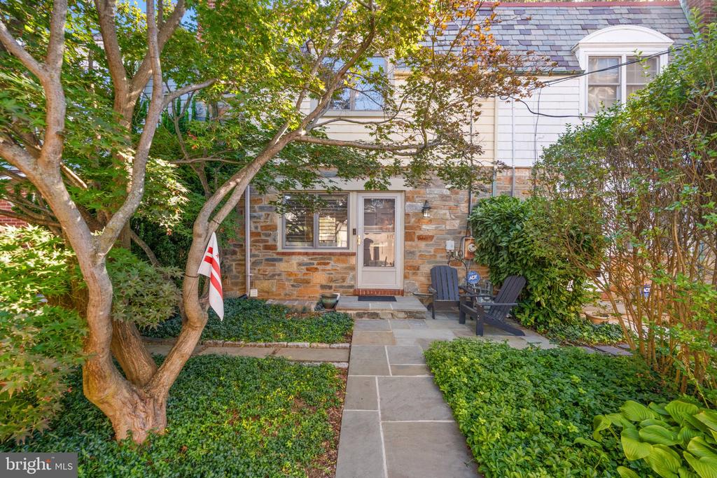 House entrance with a stone facade, garden path, and chairs beneath leafy trees.