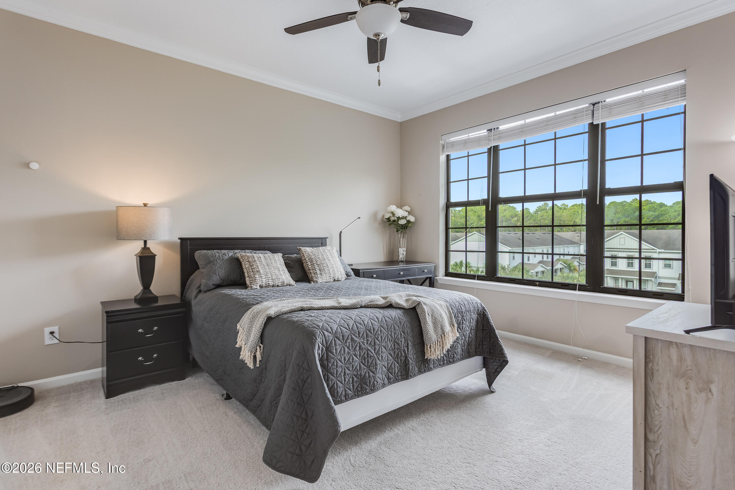 Bedroom with a gray quilted bed beside a large window overlooking homes and trees.