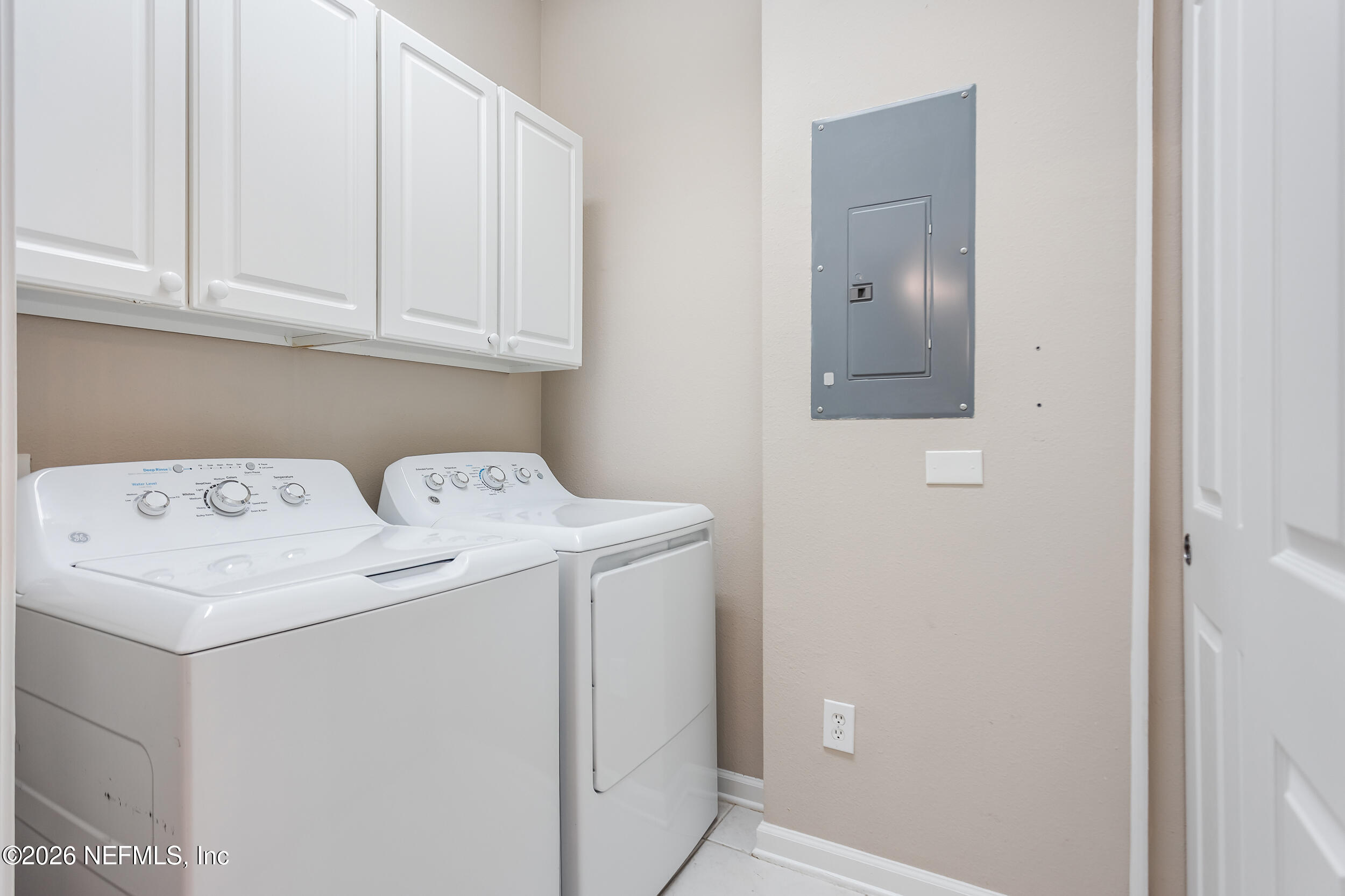 Laundry room with white washer and dryer beneath wall cabinets and an electrical panel.