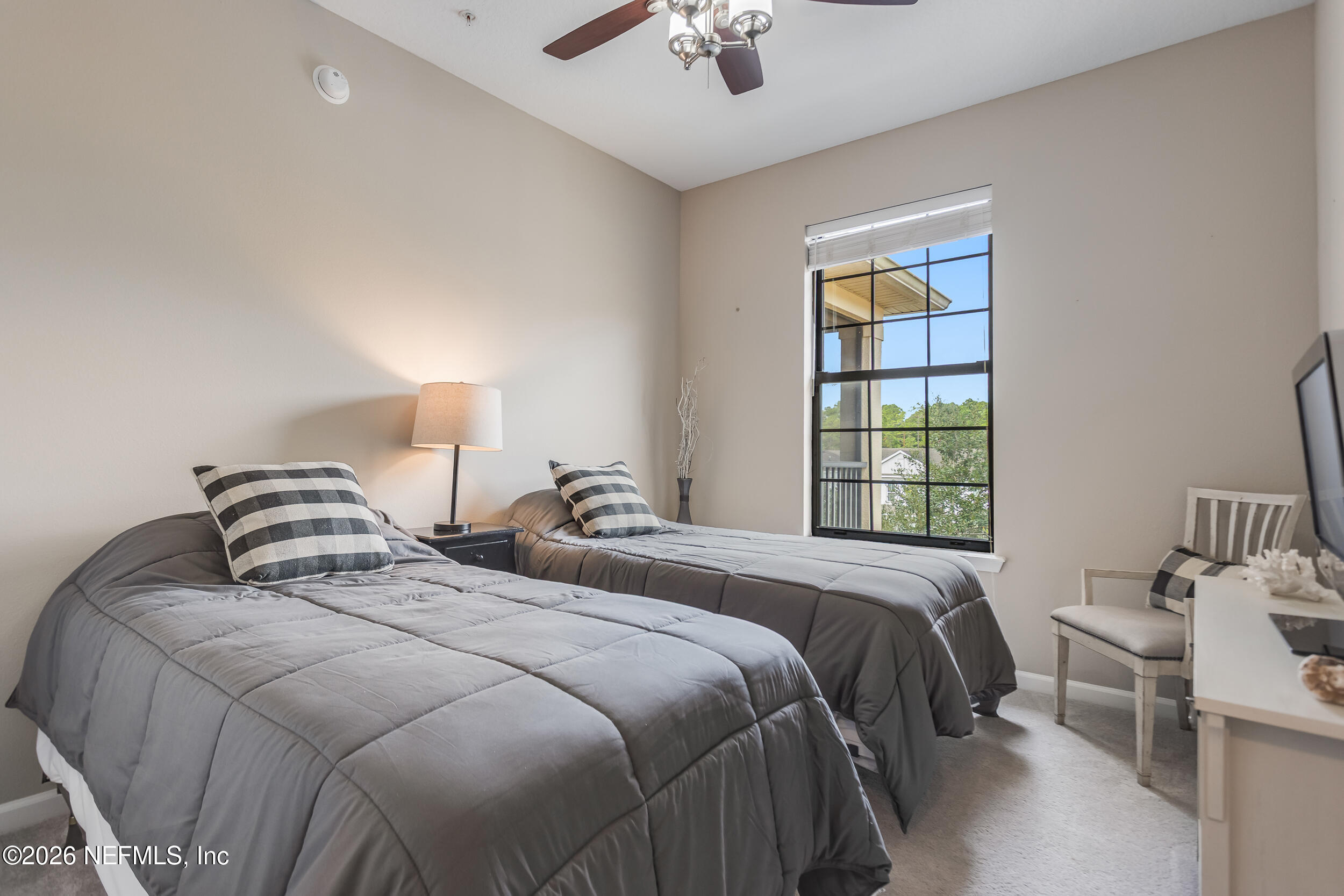 Furnished bedroom with two gray twin beds, checked pillows, and a tall window overlooking trees.