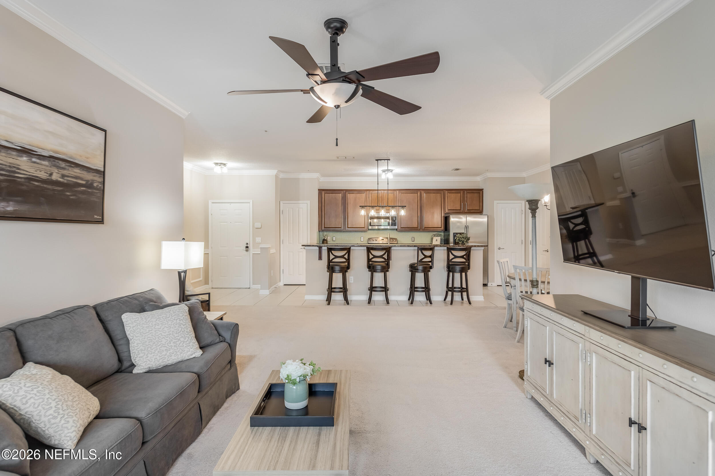 Open-plan living room and kitchen with a gray sectional, large television, and breakfast bar with four stools.