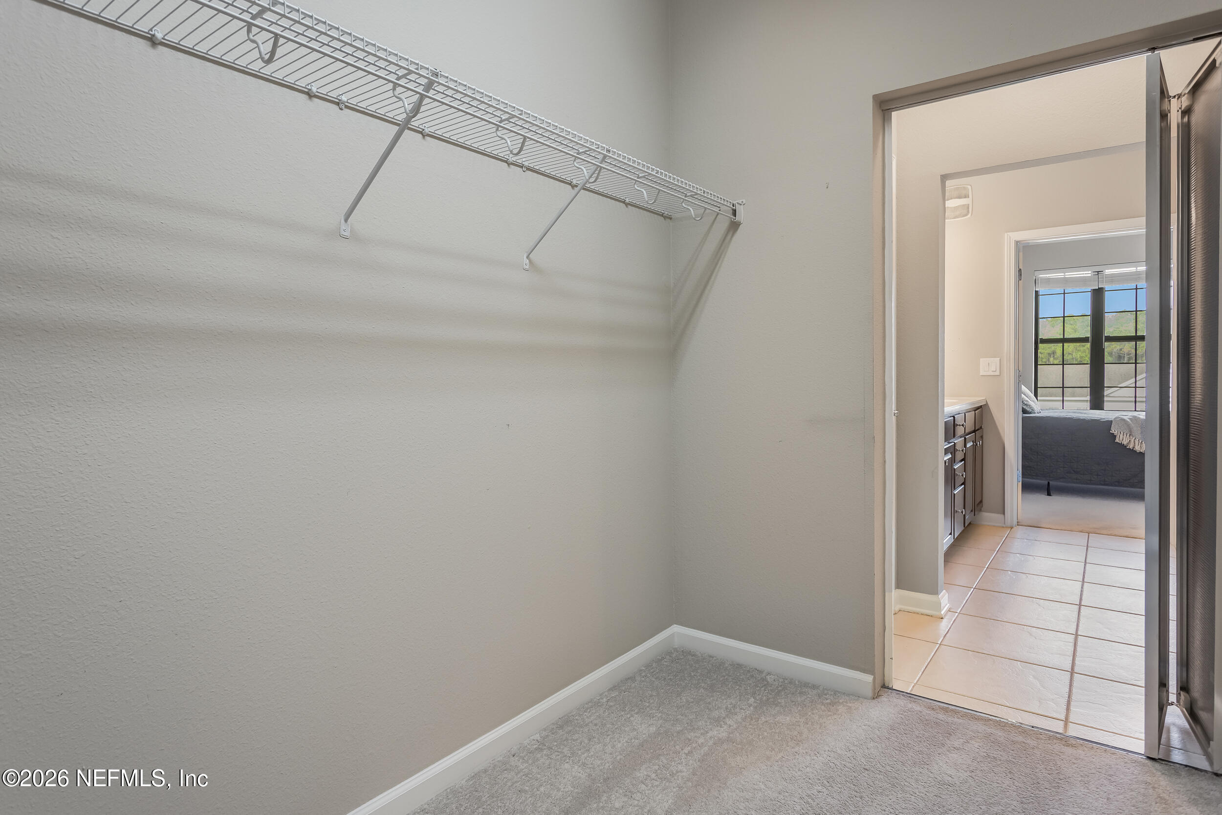 Walk-in closet with a long wire shelf, opening onto a tiled bathroom and bedroom.