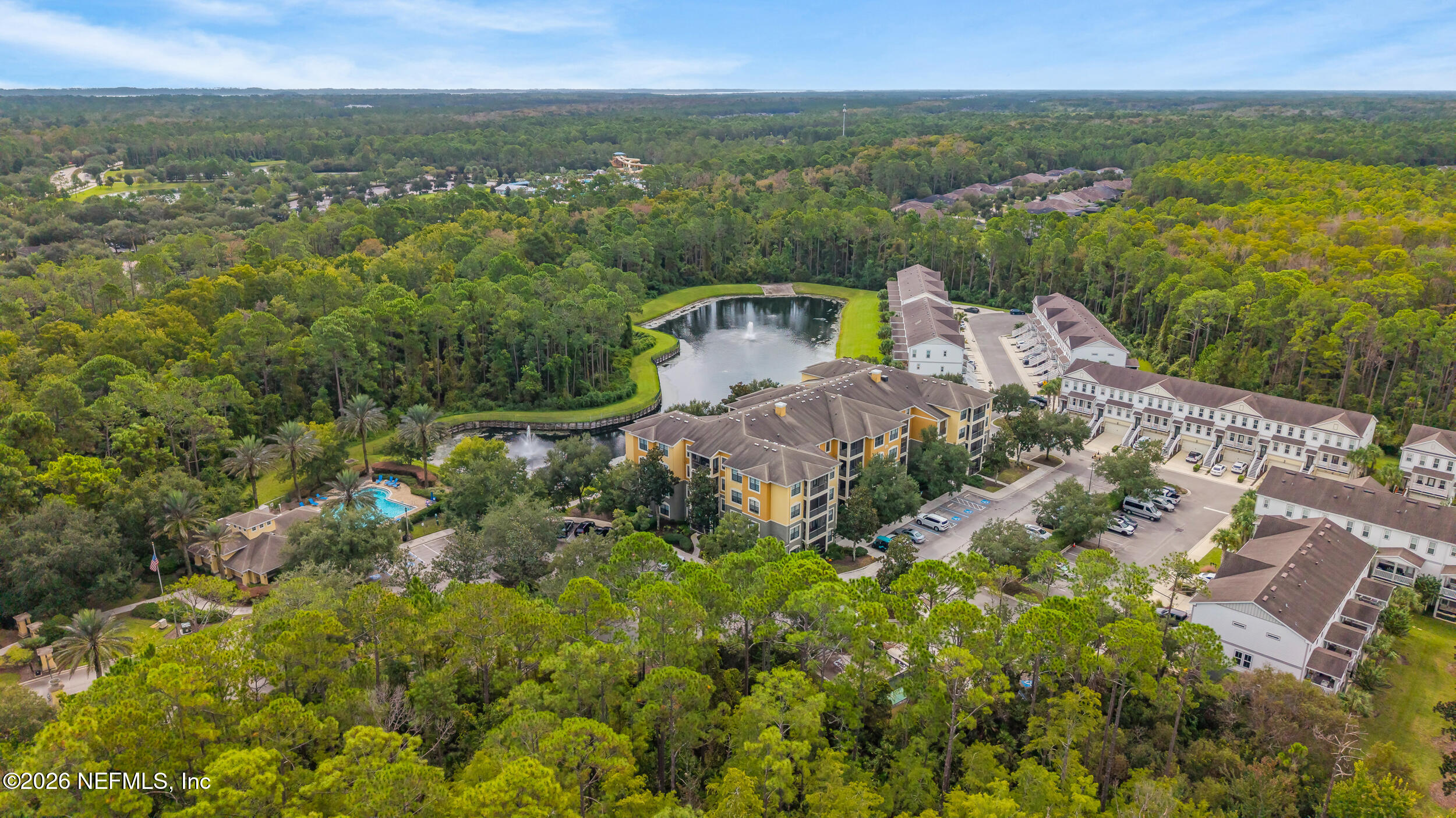 Aerial view of apartment community with a pond, fountain, swimming pool, and surrounding trees.
