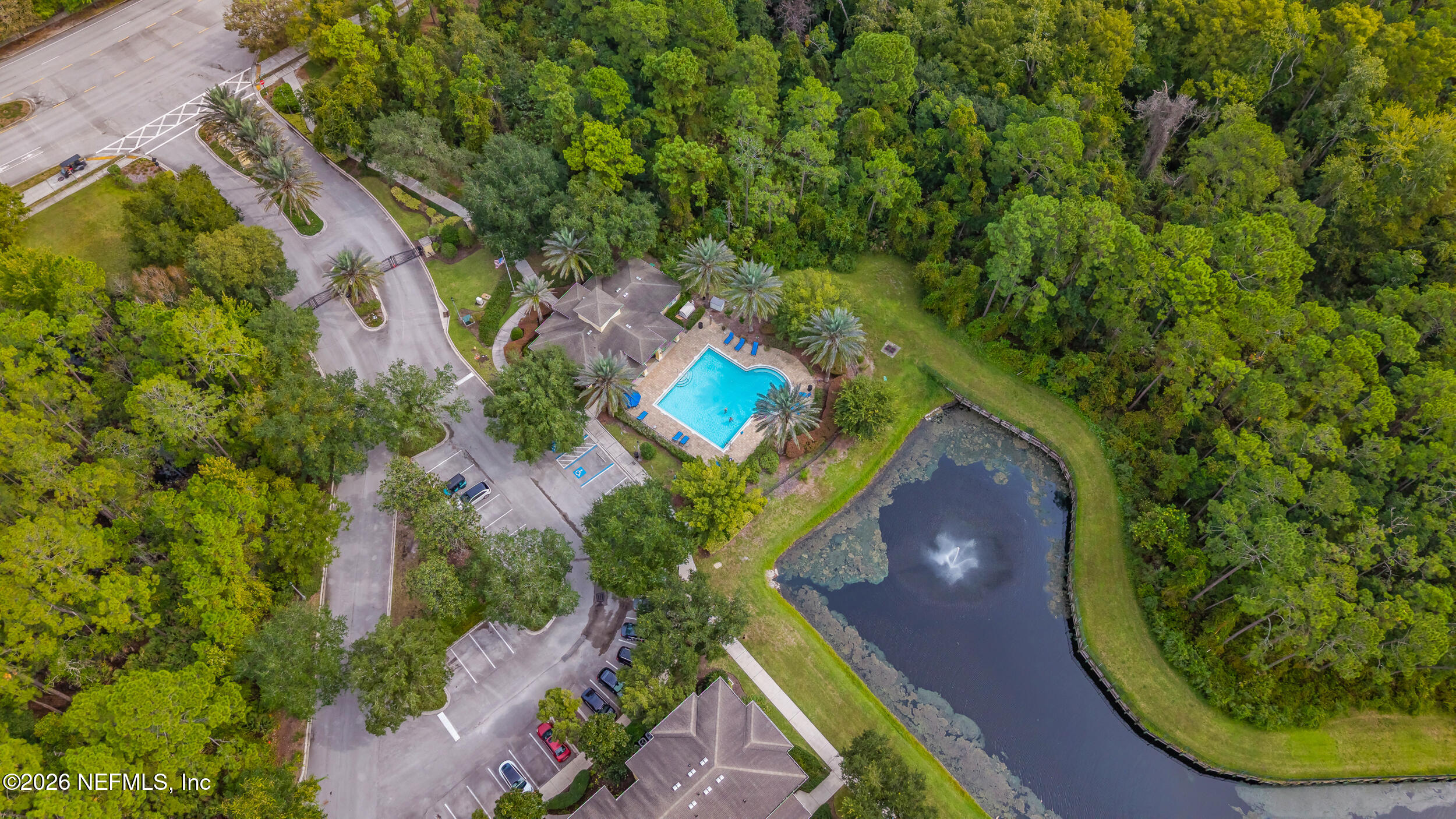Aerial view of a community pool and clubhouse beside a pond, surrounded by trees.