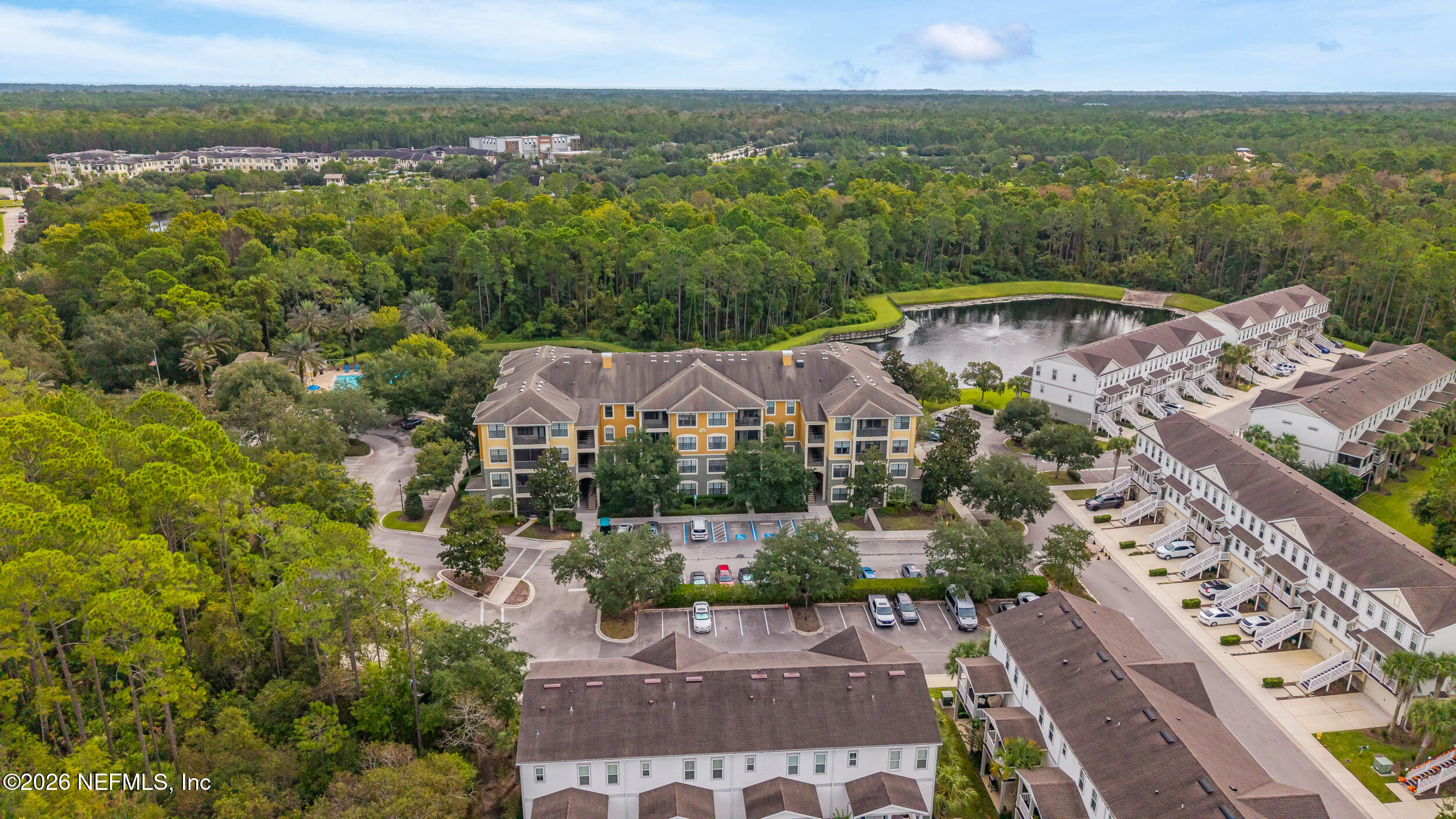 Aerial view of a residential community with apartment buildings, a pond, pool, and surrounding woods.