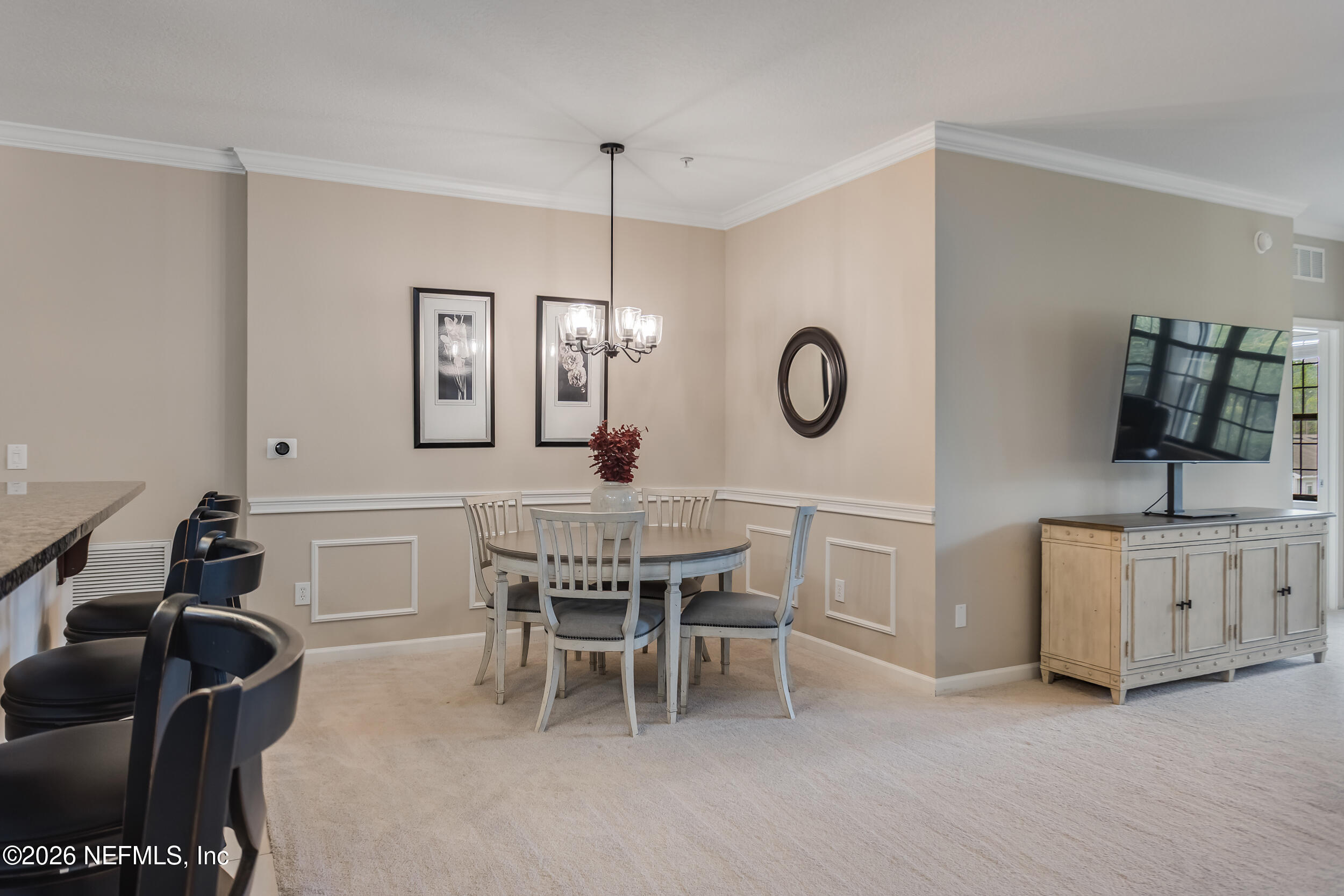 Dining area with round table and chandelier, kitchen bar stools, and adjoining living room with television.