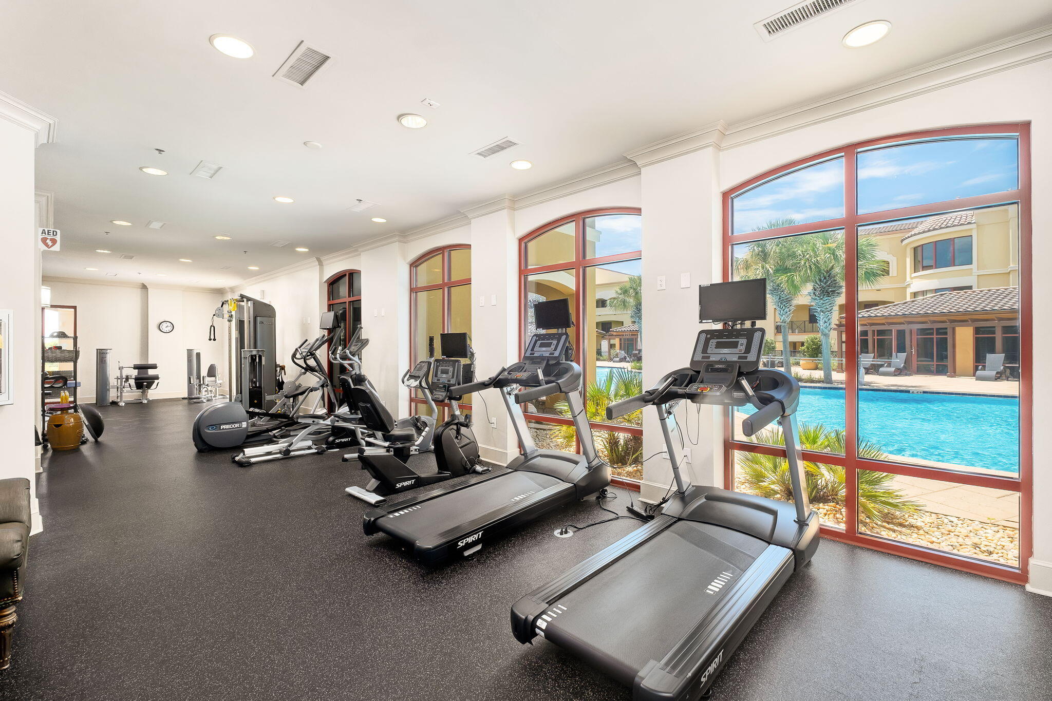 Fitness center with rows of exercise equipment and arched windows overlooking a pool.