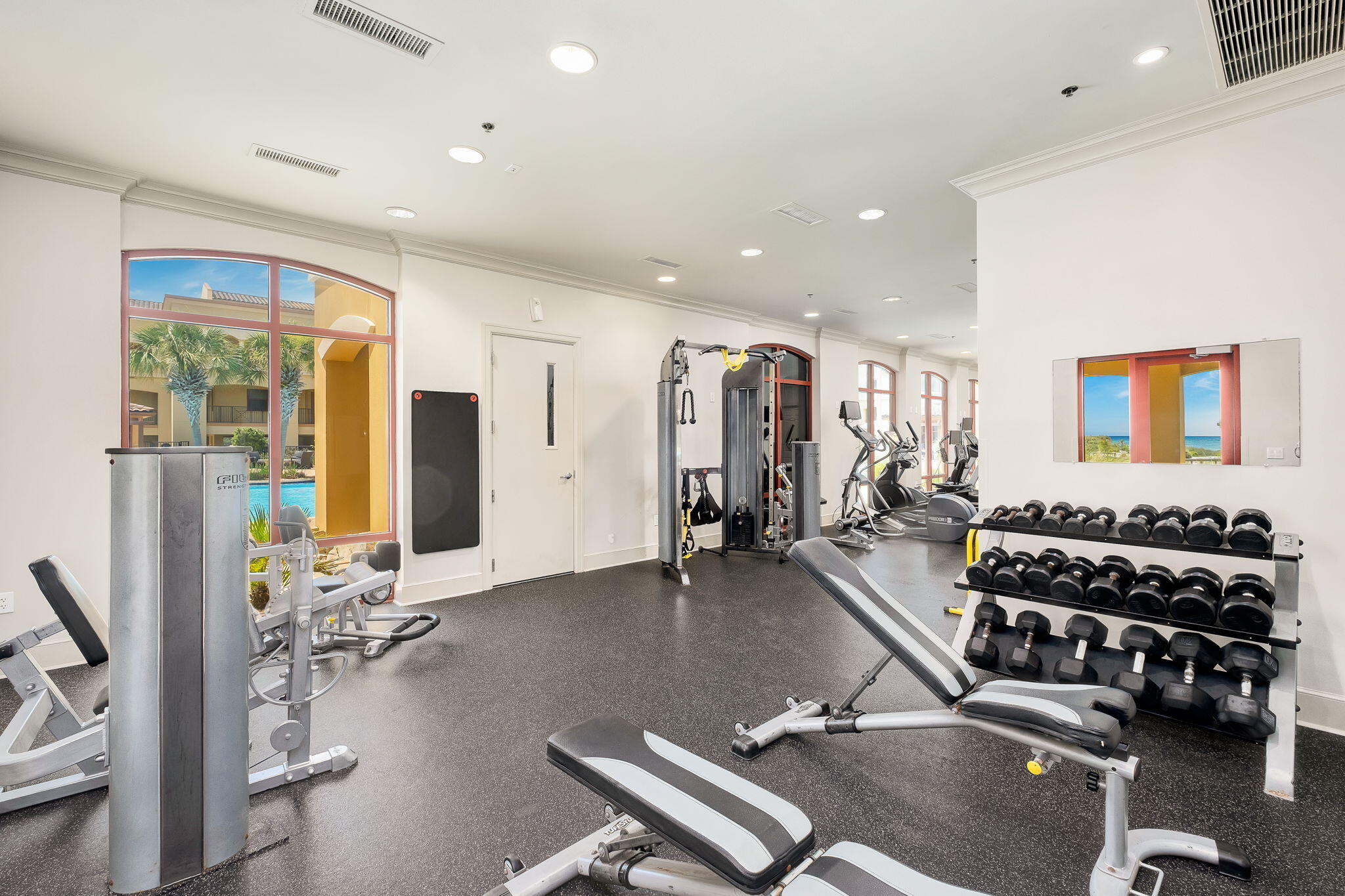 Fitness room with dumbbell racks, weight machines, and arched windows overlooking a pool courtyard.