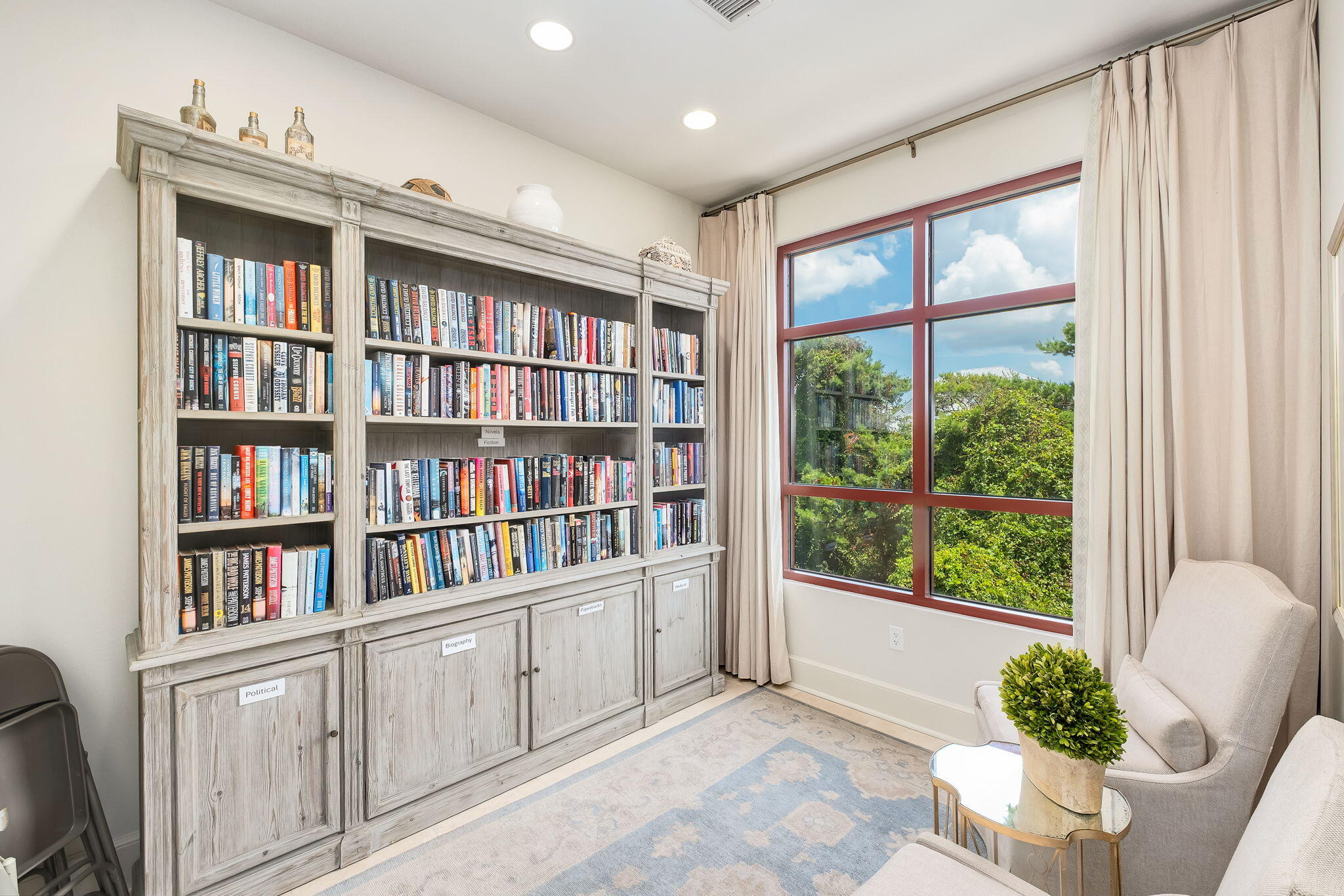 Furnished library with a wall of bookshelves beside a large window overlooking green trees.