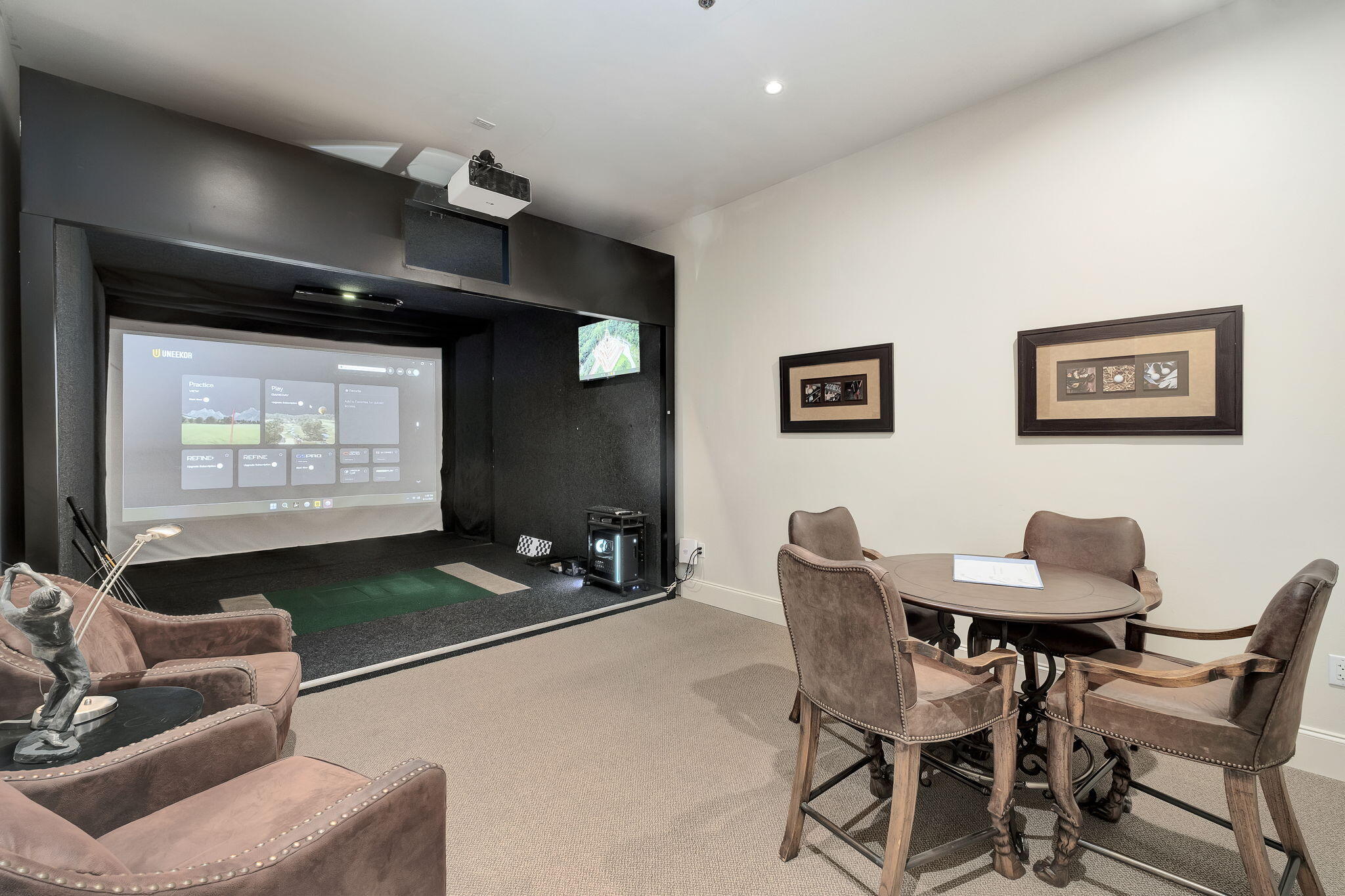Golf simulator room with a large projection screen, green hitting area, and seating around a table.