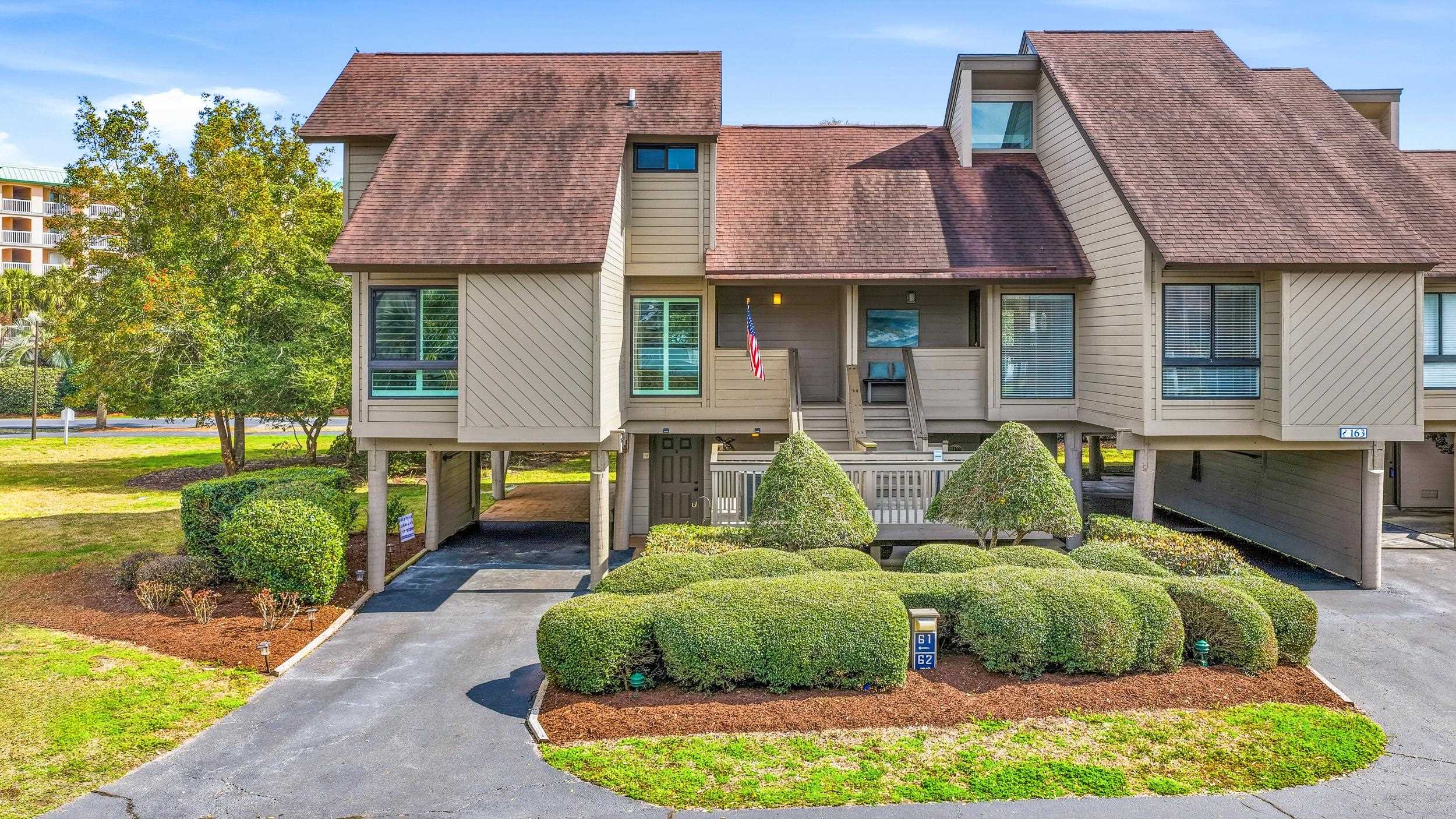 Elevated beige townhouse with a brown shingle roof, covered porch, and landscaped front entry.