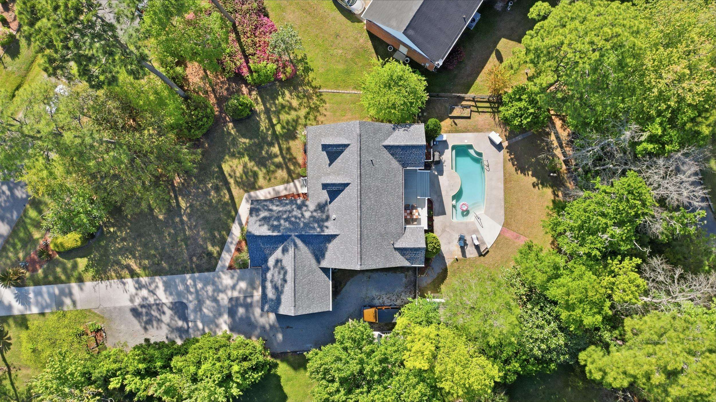Aerial view of a home with a swimming pool, broad driveway, and tree-filled landscaped yard.