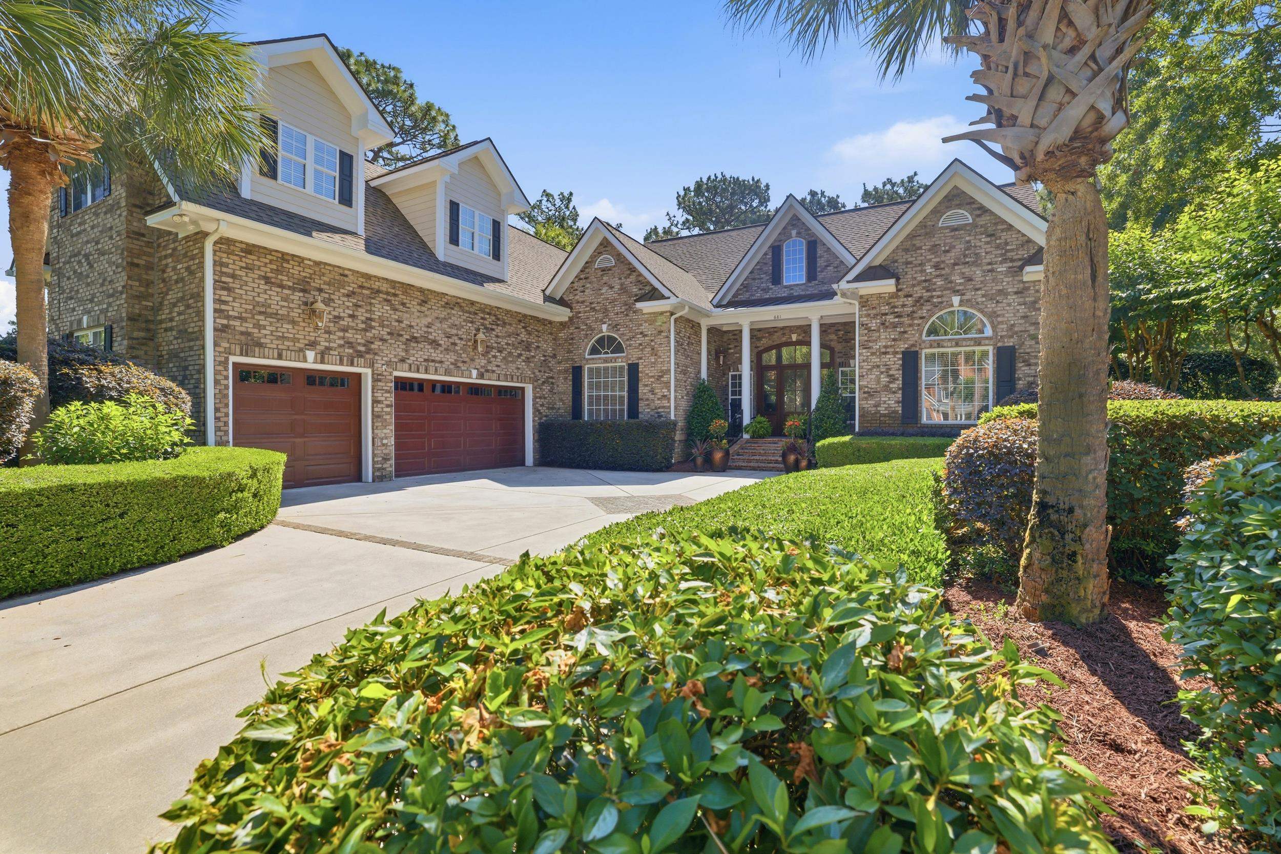 Large brick home with two garage doors, a broad driveway, trimmed hedges, and palm trees.