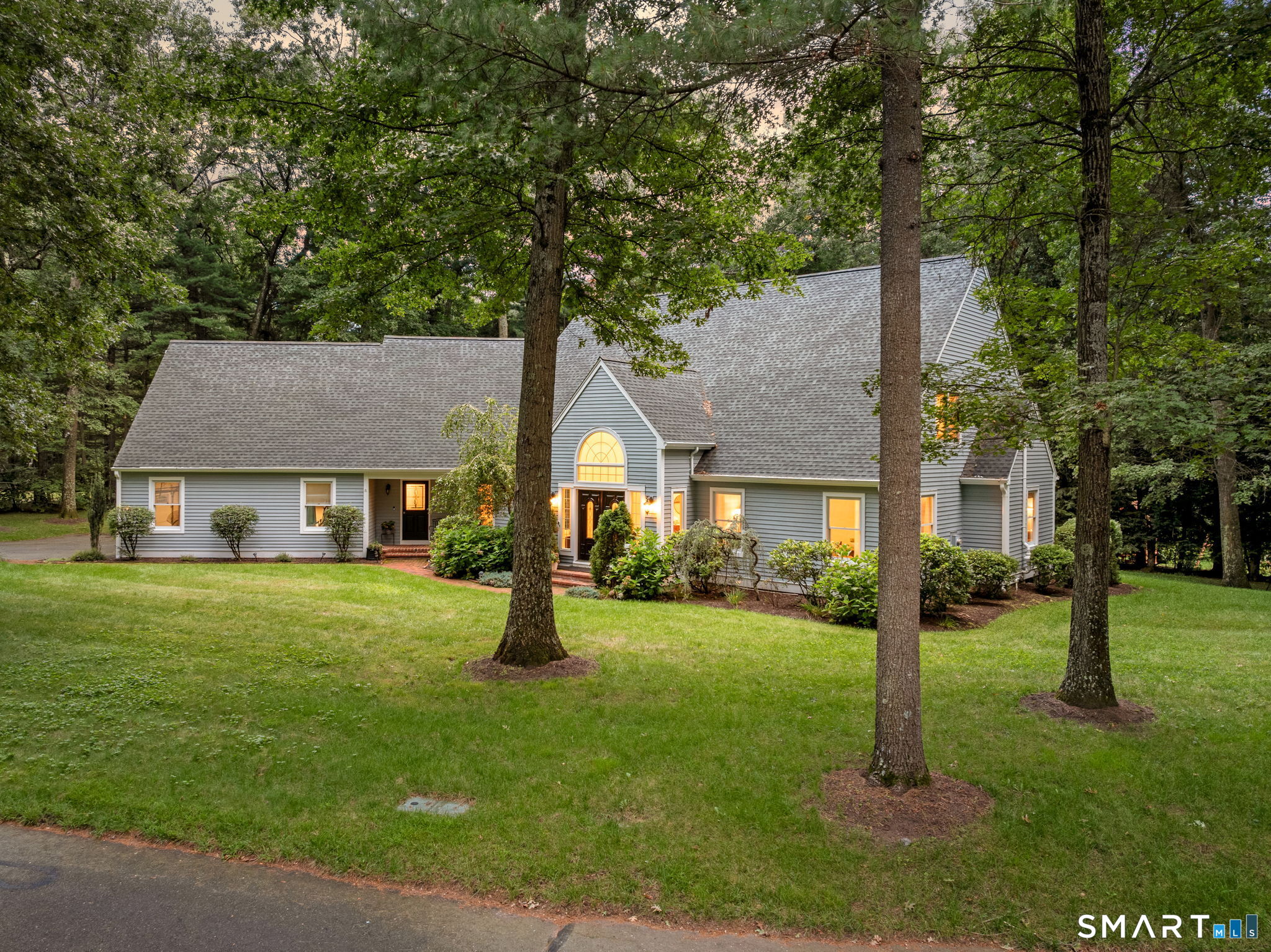 Gray-sided house with an arched entry window, landscaped lawn, and mature trees.