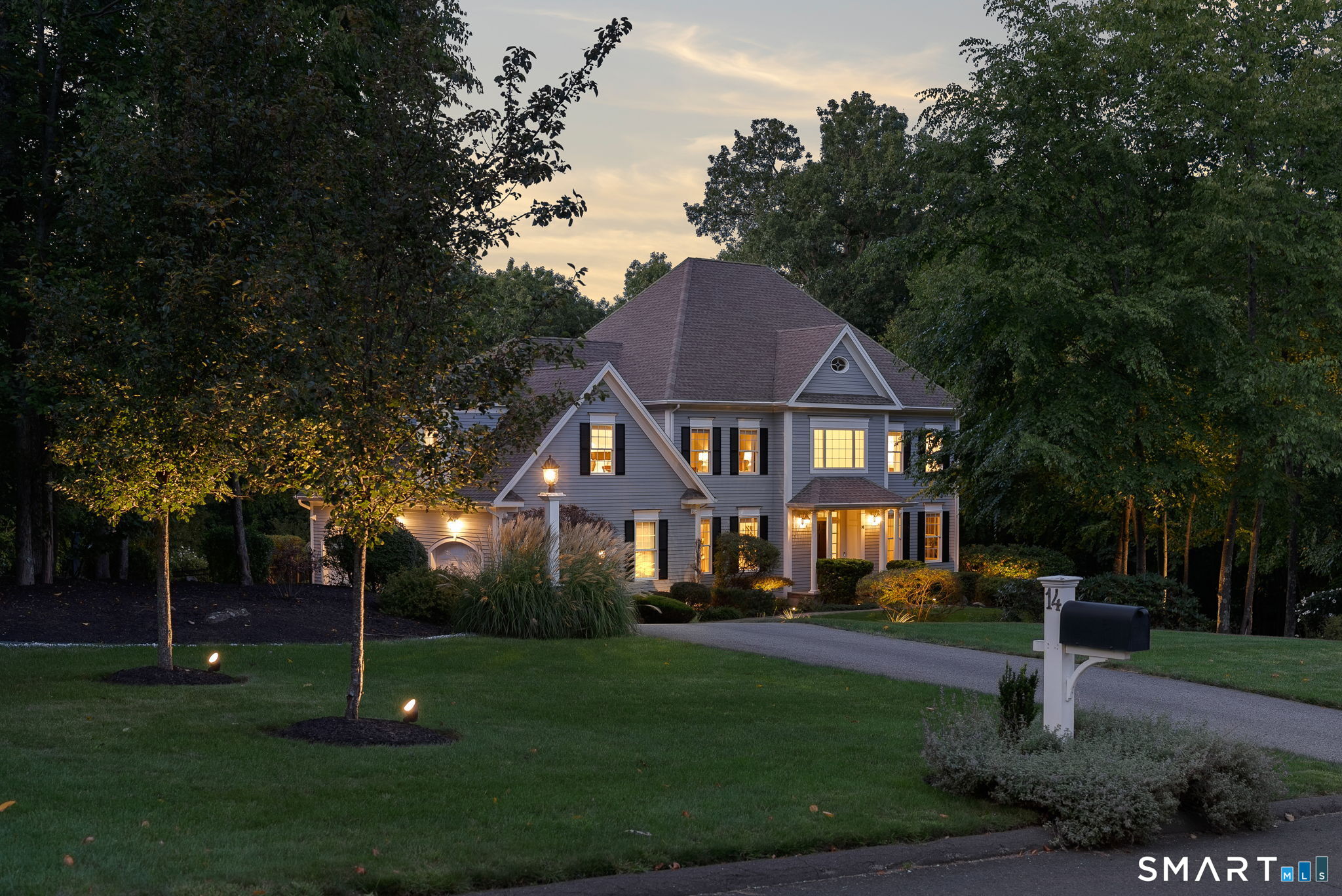 Gray two-story home at dusk, with warm window lights, mature trees, and a landscaped lawn.