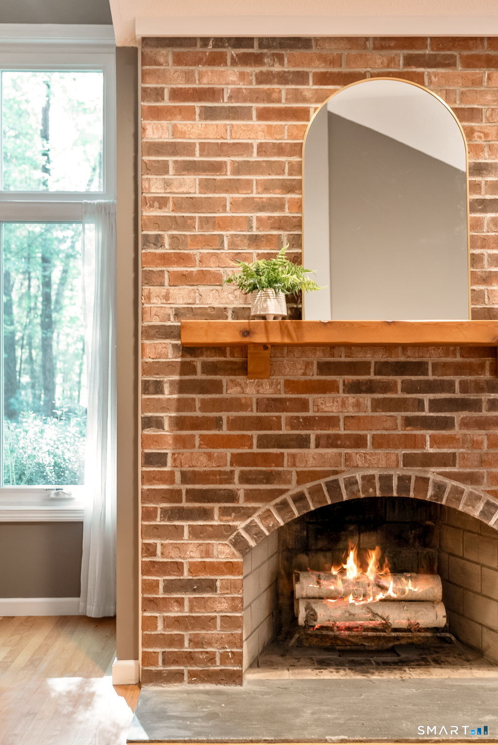 Brick fireplace with a burning fire, wood mantel, arched mirror, and fern beside a curtained window.