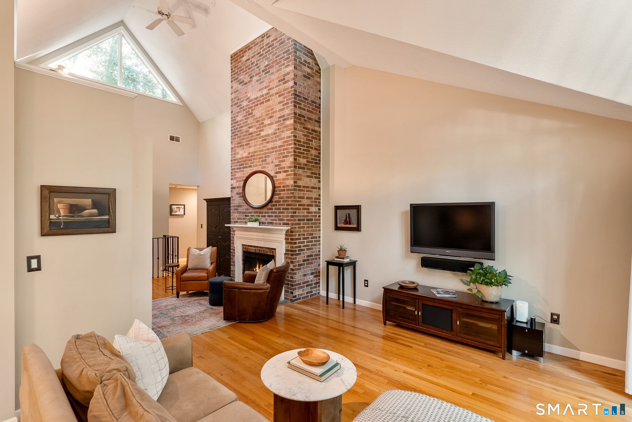 Living room with vaulted ceiling, brick fireplace, triangular window, and hardwood floors