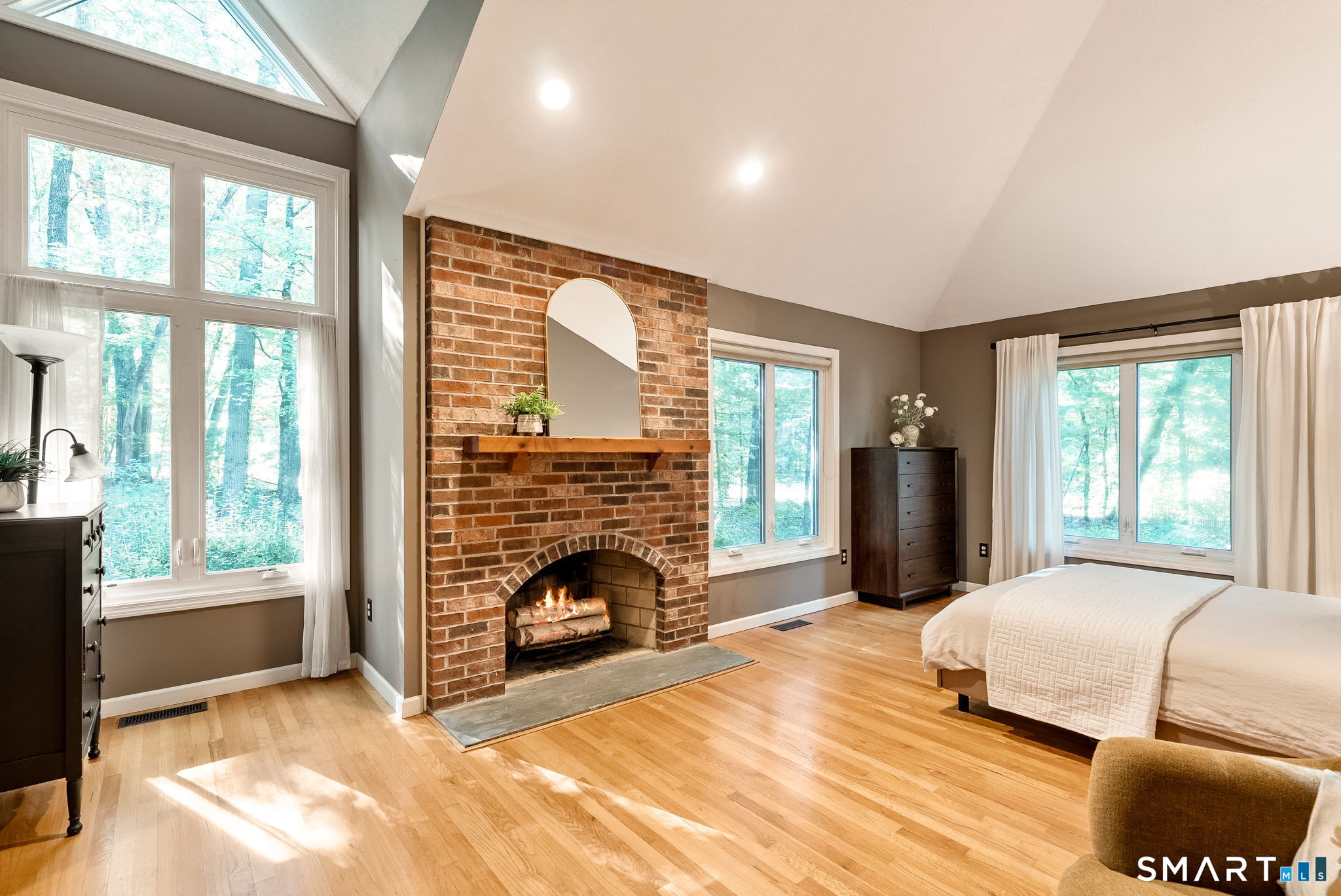 Bedroom with a brick fireplace, vaulted ceiling, skylight, and large windows overlooking trees.