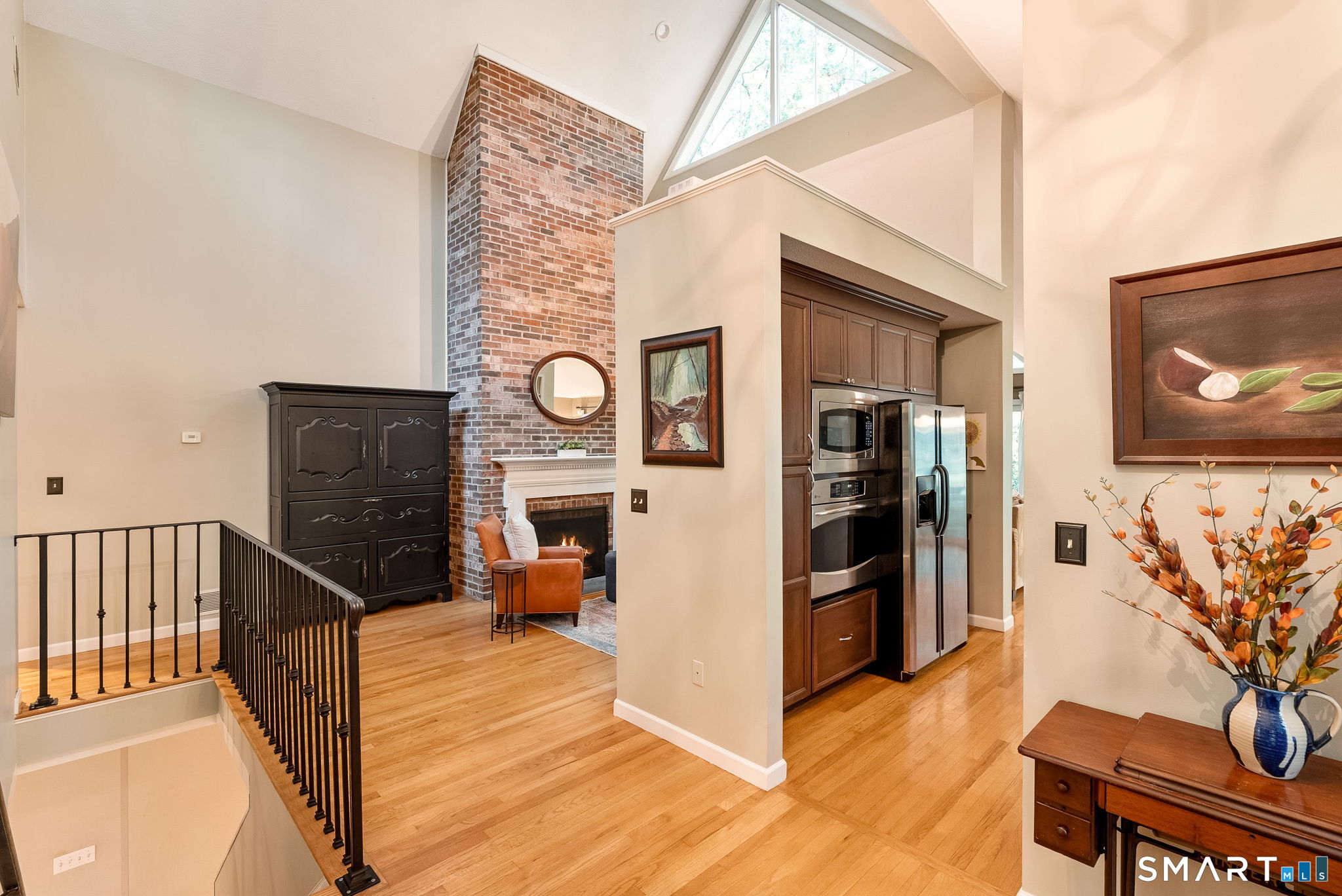 Open-plan living area with a tall brick fireplace, vaulted ceiling, hardwood floors, and adjoining kitchen.