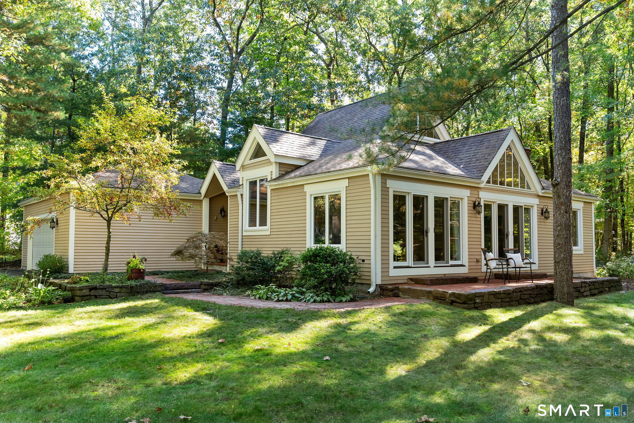 Beige house with white trim and a brick patio surrounded by mature trees.