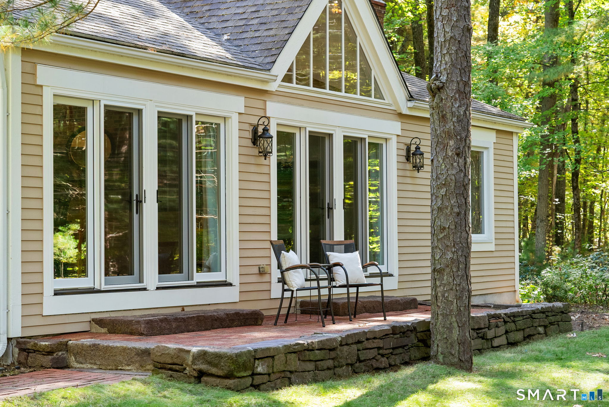 Beige home with tall windows and a raised stone patio with two chairs among mature trees.