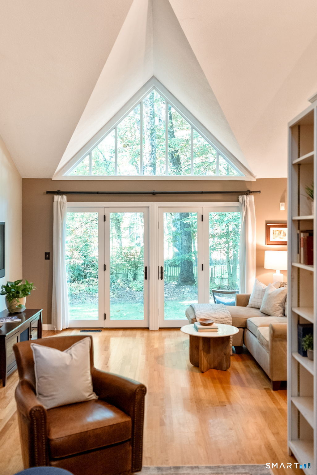 Living room with a vaulted ceiling, triangular window, and French doors overlooking trees.