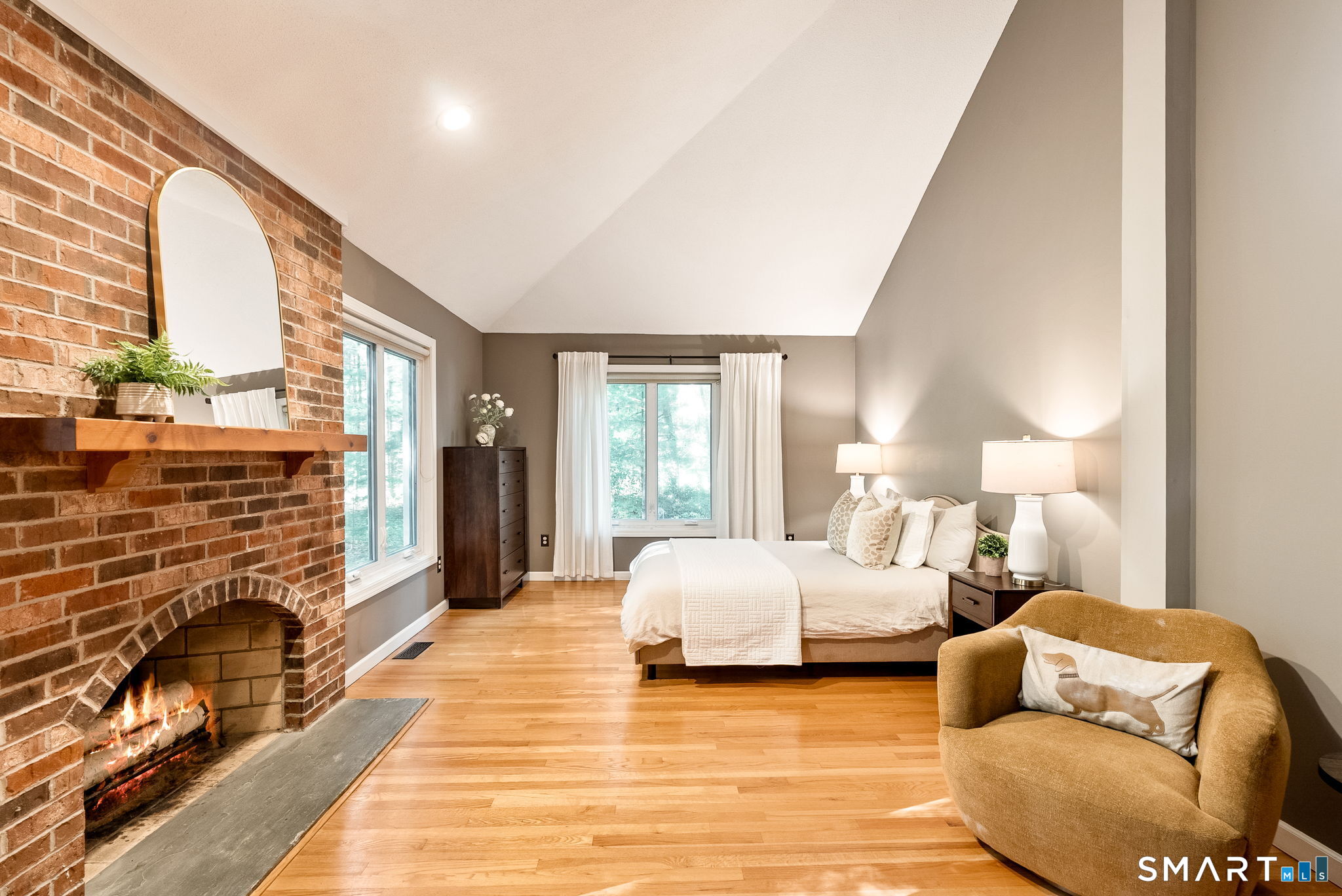 Staged bedroom with a brick fireplace, sloped ceiling, hardwood floors, and an upholstered chair.