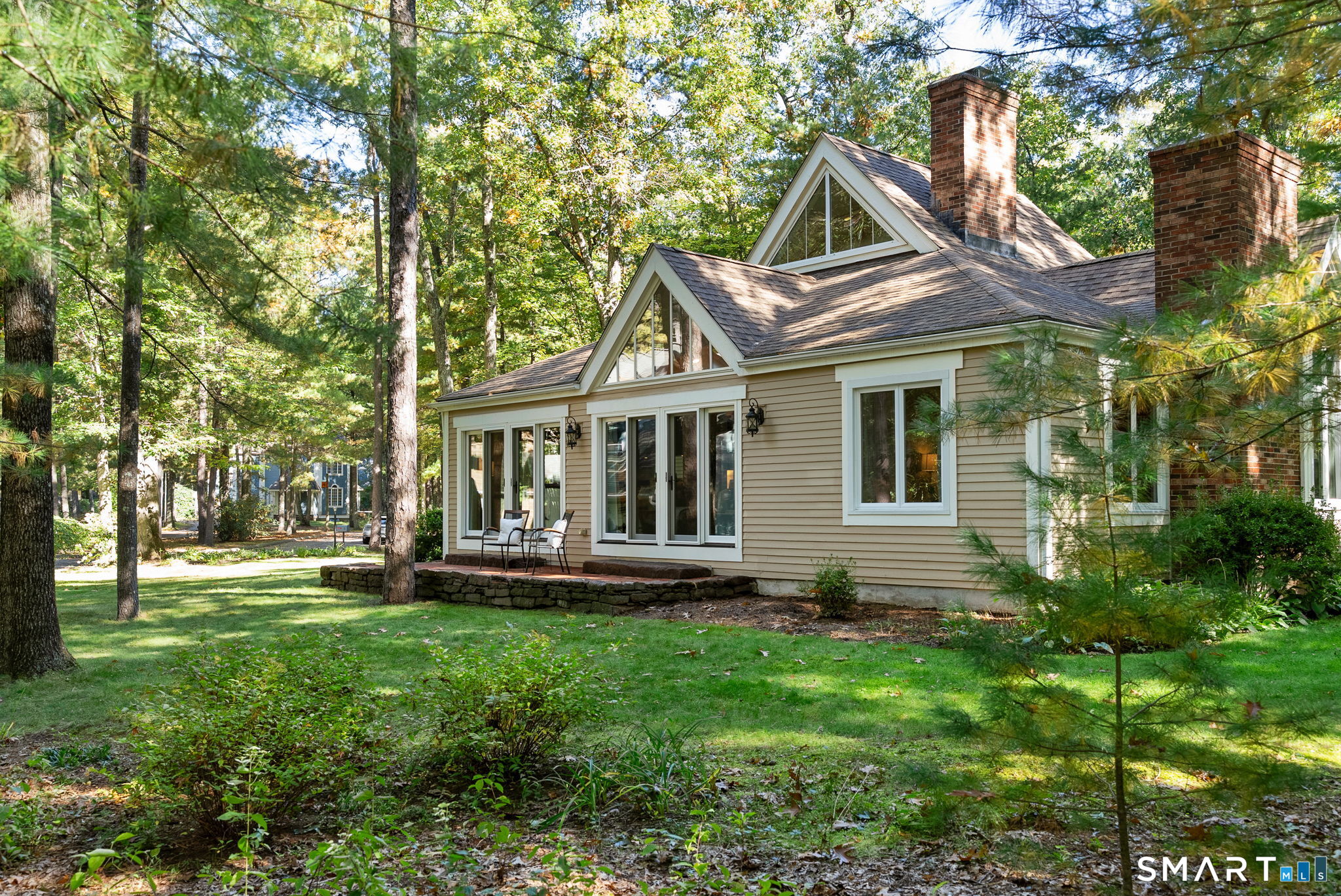 House rear with gabled roof and brick chimneys, set among mature trees beside a stone patio.