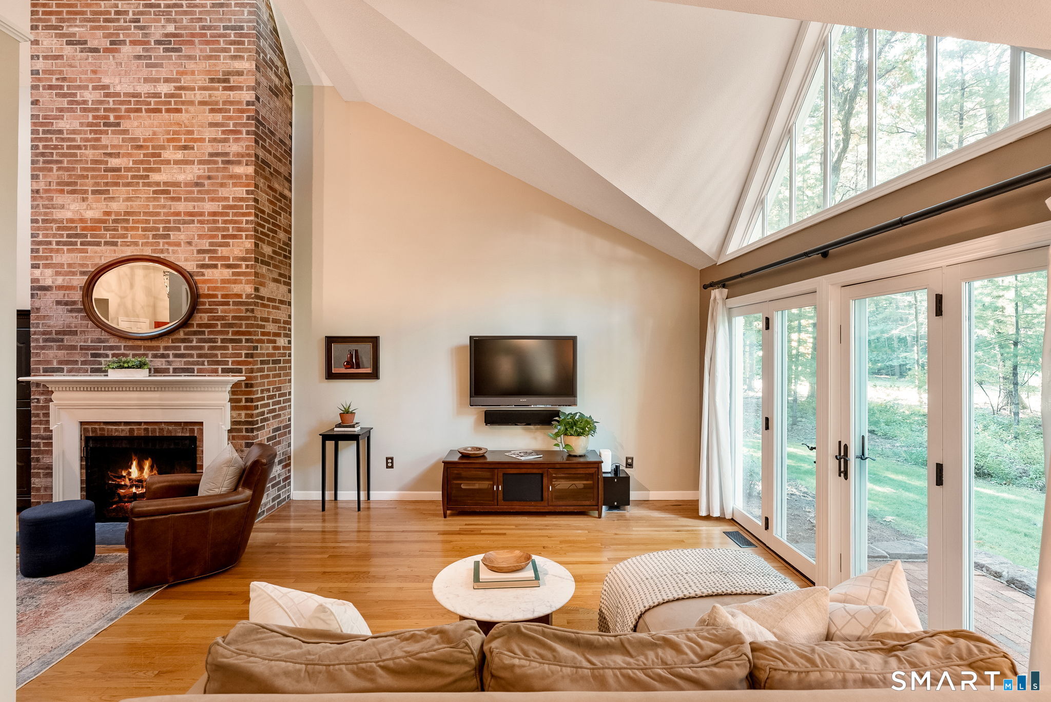 Living room with a brick fireplace, vaulted ceiling, and glass doors overlooking trees.