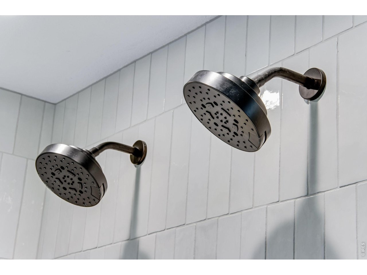 Two dark metal showerheads mounted on a white tiled bathroom wall.
