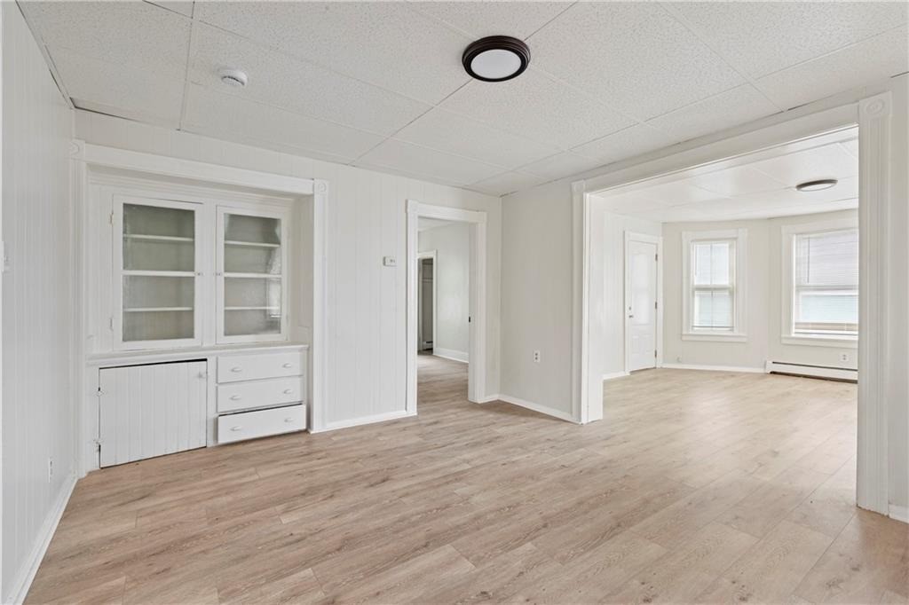 Empty interior with built-in white cabinetry and light wood-look floors opening into a bright room with two windows.