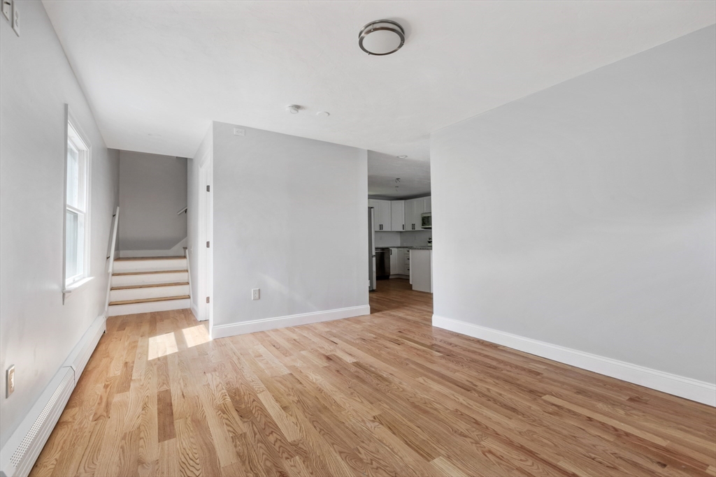 Vacant living area with light hardwood floors, a staircase, and a glimpse of the kitchen.