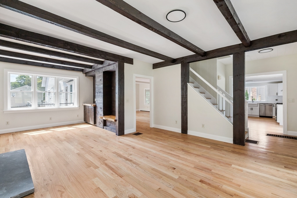 Vacant living room with exposed dark beams, hardwood floors, and a staircase beside the kitchen.