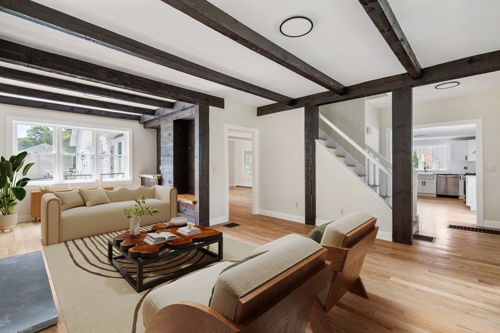 Living room with exposed dark ceiling beams, hardwood floors, and adjoining staircase and kitchen.