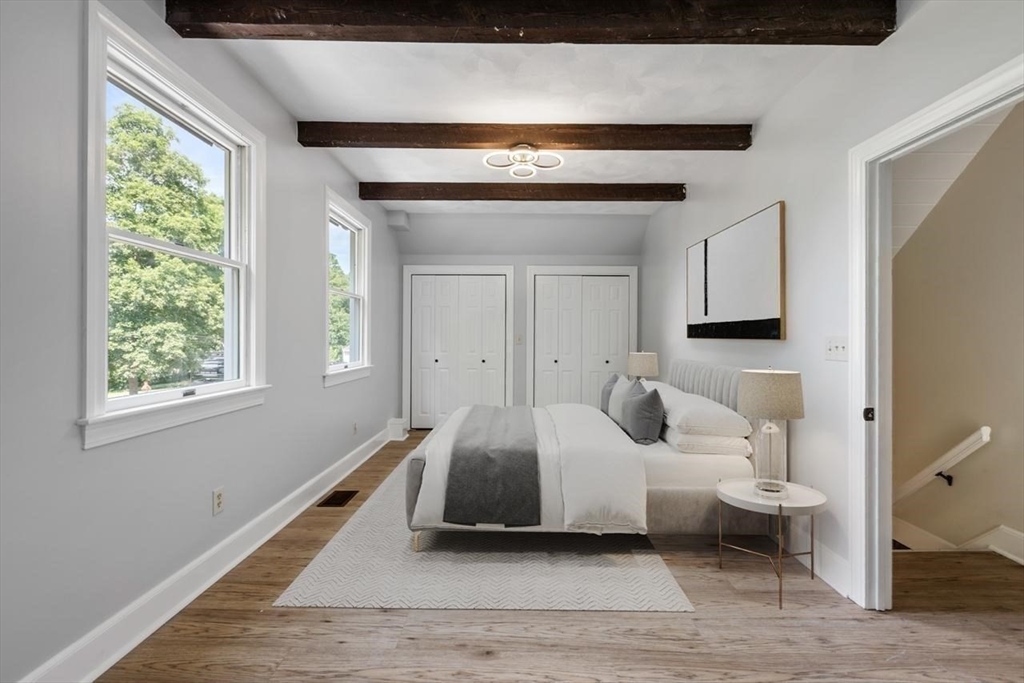 Staged bedroom with a white bed, exposed dark ceiling beams, and large windows.