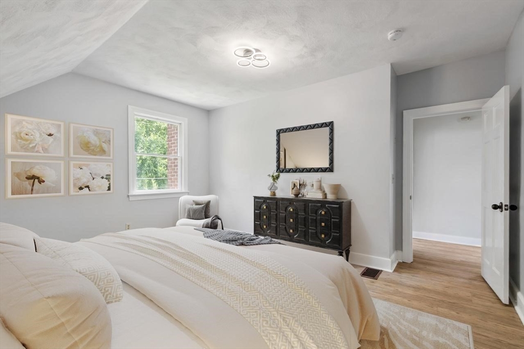 Staged bedroom with cream bedding, floral wall art, and a dark dresser beneath a mirror.
