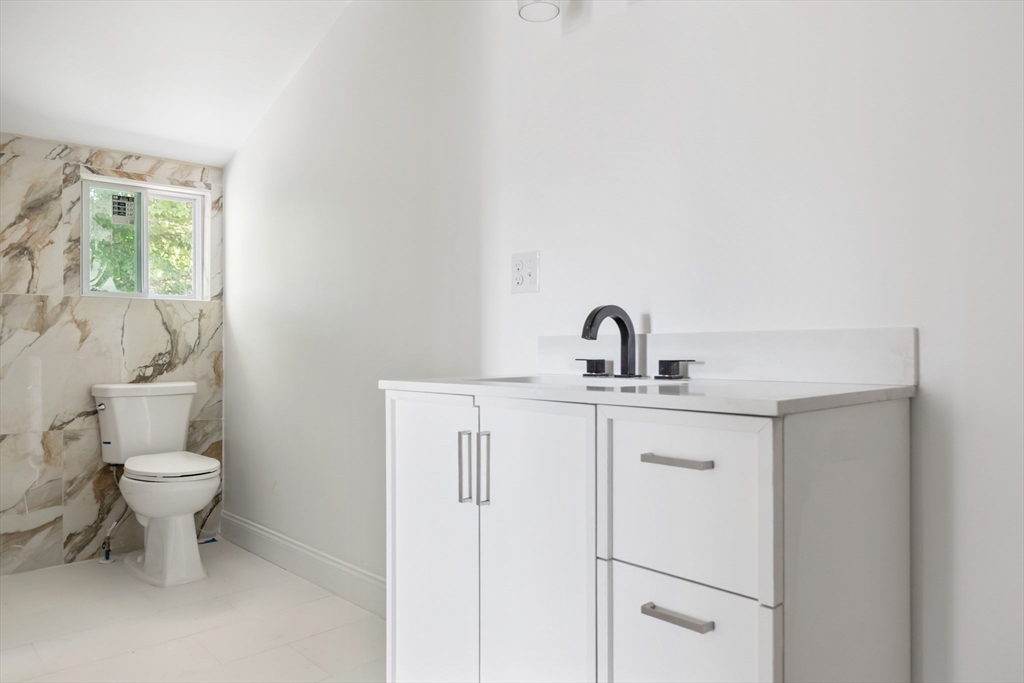 Bathroom with a white vanity and black faucet, marble-look tile, toilet, and small window.