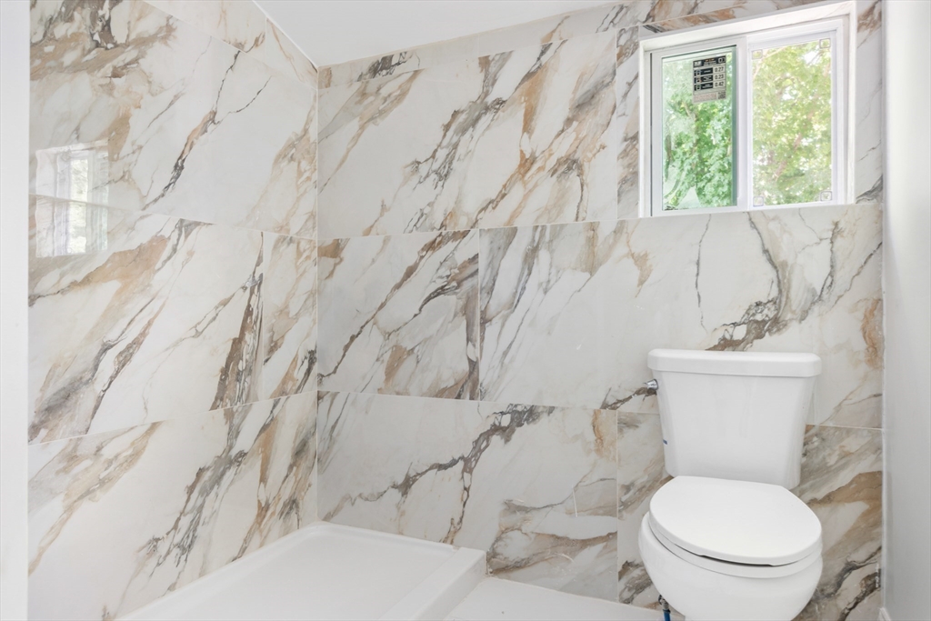Bathroom with a white toilet, shower base, and dramatic marble-look tiled walls beside a small window.