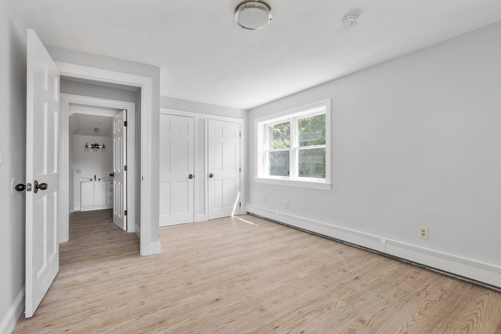 Empty bedroom with light wood flooring, white closet doors, and a window beside an open bathroom doorway.