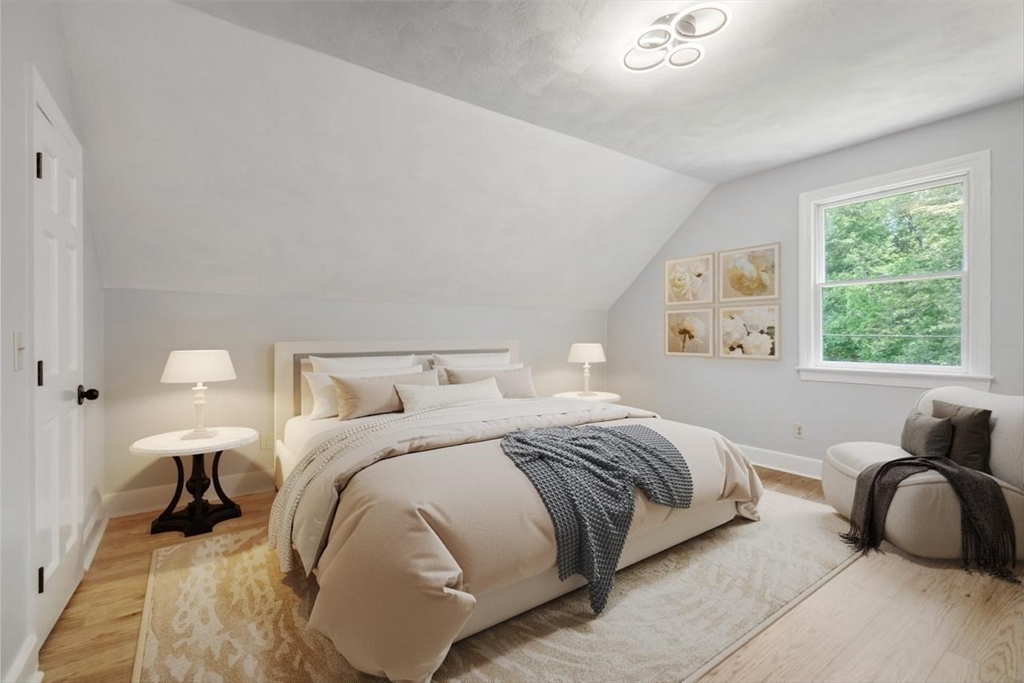 Staged bedroom with a cream bed, gray throw, sloped ceiling, and window overlooking trees.