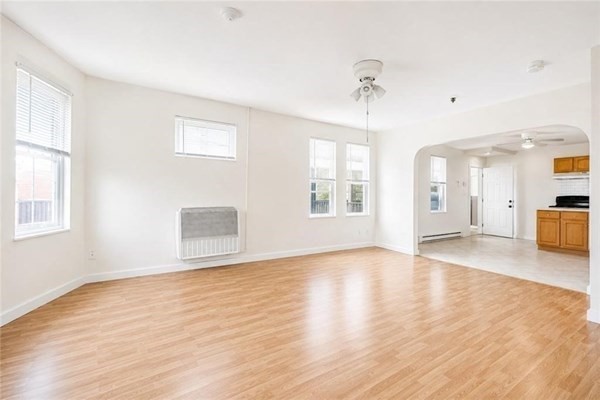 Vacant living area with light wood floors, several windows, and an arched opening to the kitchen.